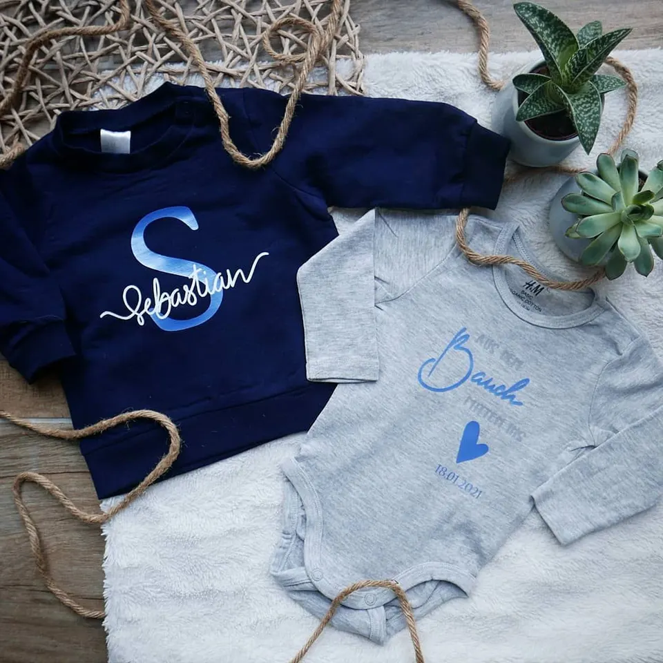 Navyblauer Kinderpulli mit S und Sebastian, daneben ein Body mit Daniel