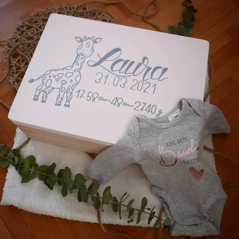 Weiße Erinnerungsbox mit Giraffe und dem Namen Laura, daneben ein Babybody