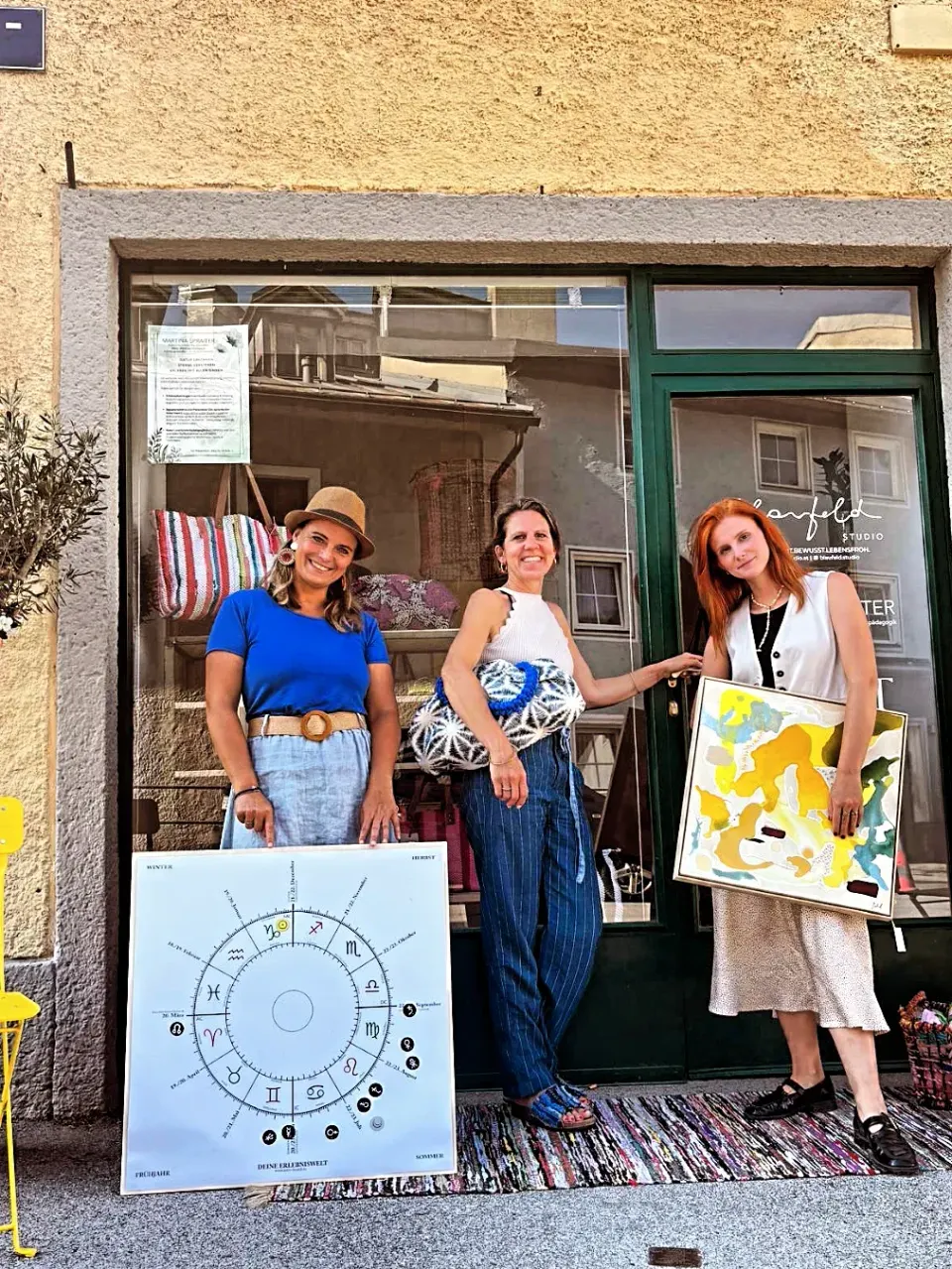 Drei Frauen vor der grünen Glasfront des Studios in der Metzgergasse, mit einer Astrologie-Tafel, einer gewebten Tasche und einem Gemälde